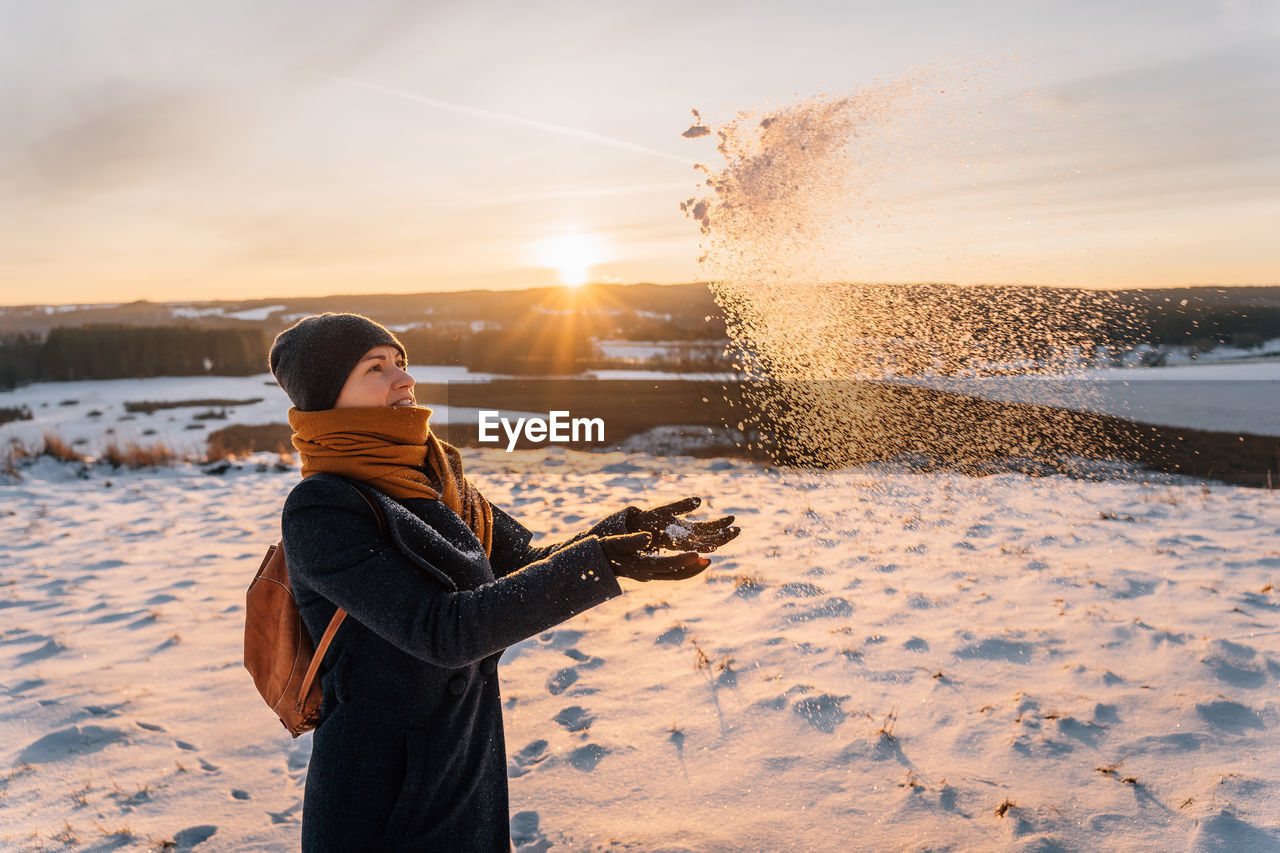 A woman in winter in warm clothes throws her palms and snow against the background of sunset.