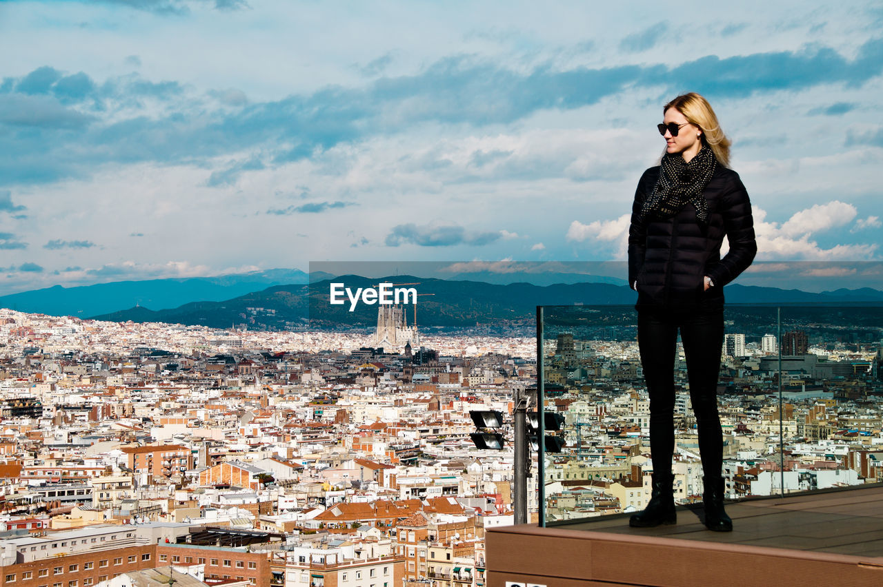 Young woman wearing warm clothing while standing on balcony against cityscape