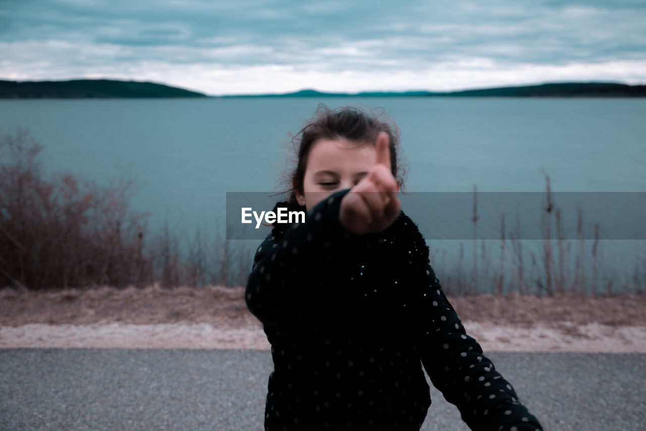 Girl pointing while standing on road by lake against cloudy sky during sunset