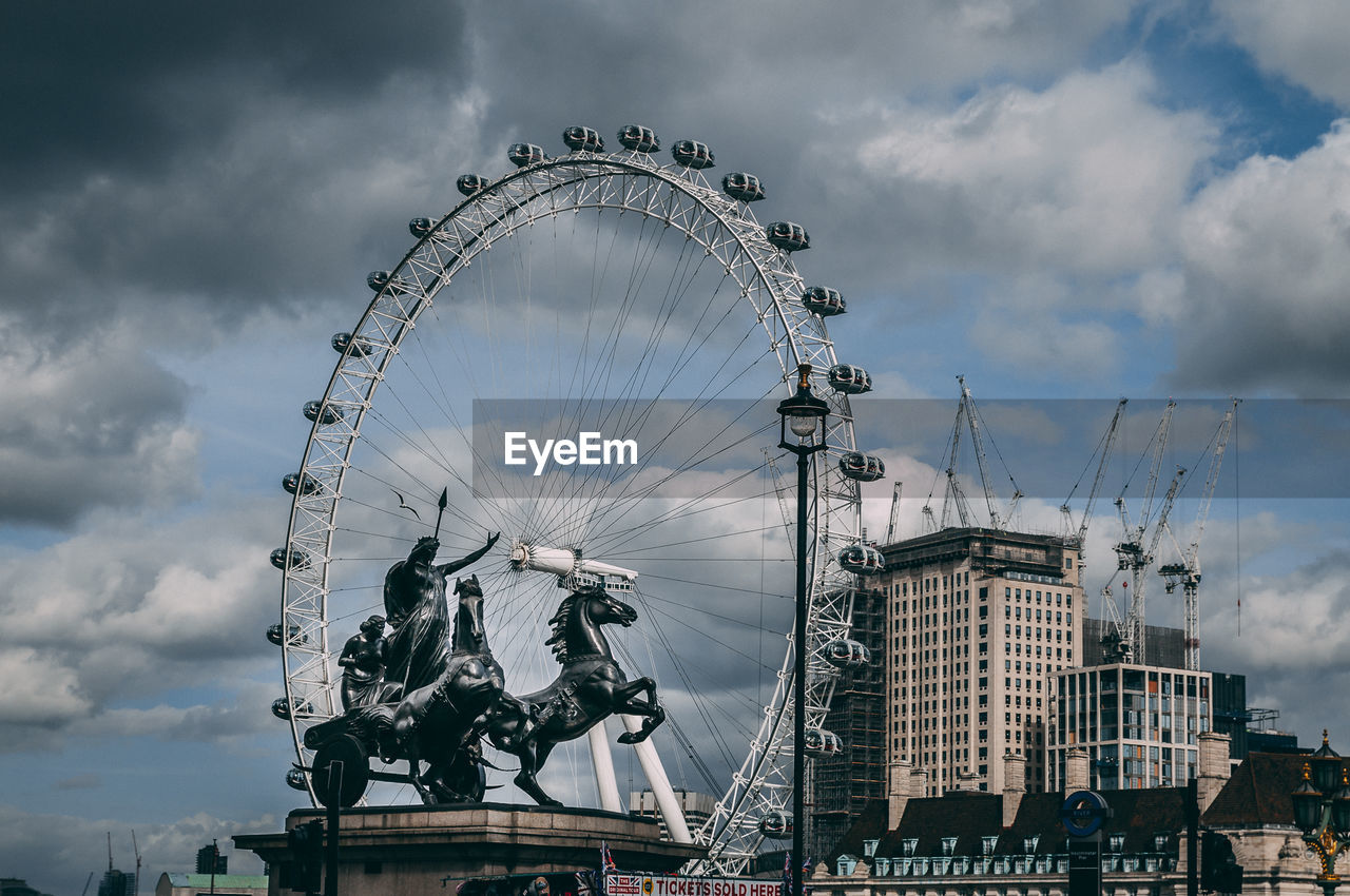 Statues and ferris wheel in city against cloudy sky