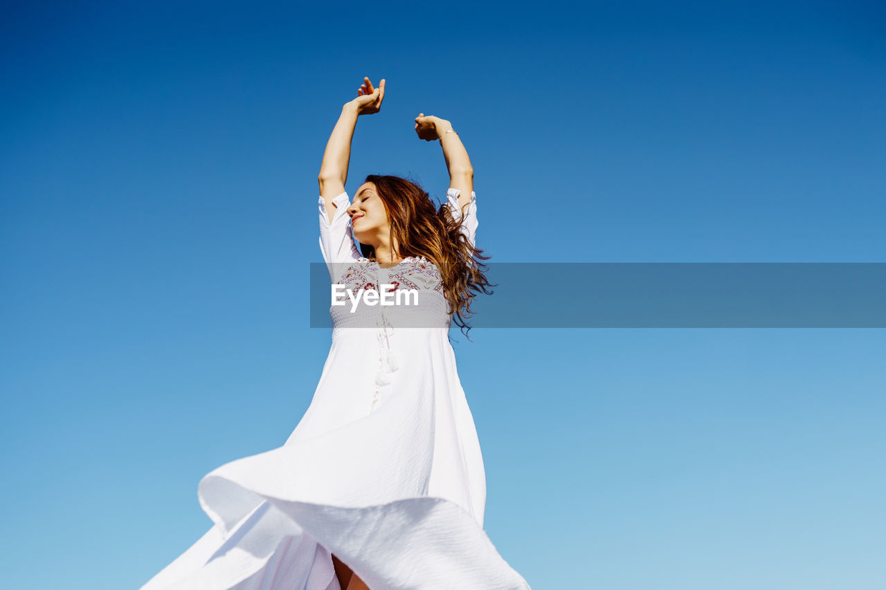 Low angel view of woman with arms raised against clear blue sky