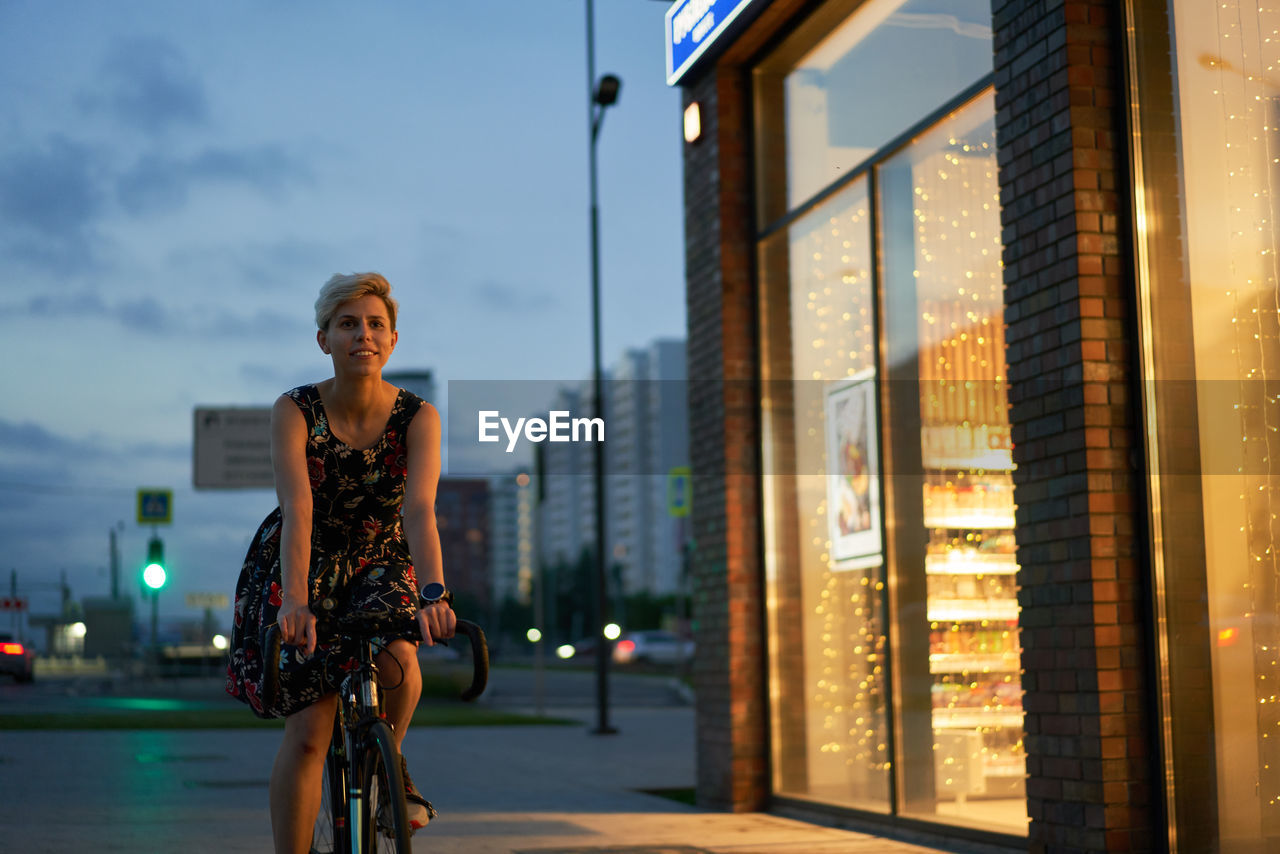 Woman in dress rides bicycle in night city near store