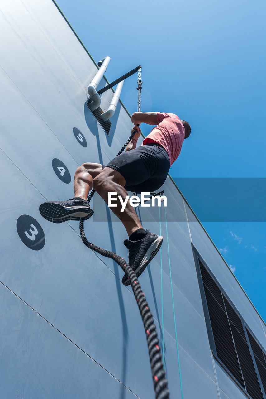 From below of confident male athlete climbing rope while doing exercises on sports ground in summer on sunny day