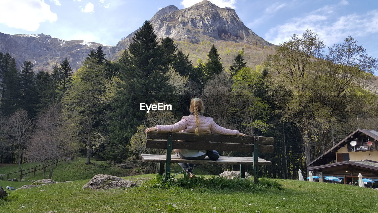 REAR VIEW OF MAN SITTING ON BENCH IN FIELD AGAINST MOUNTAIN