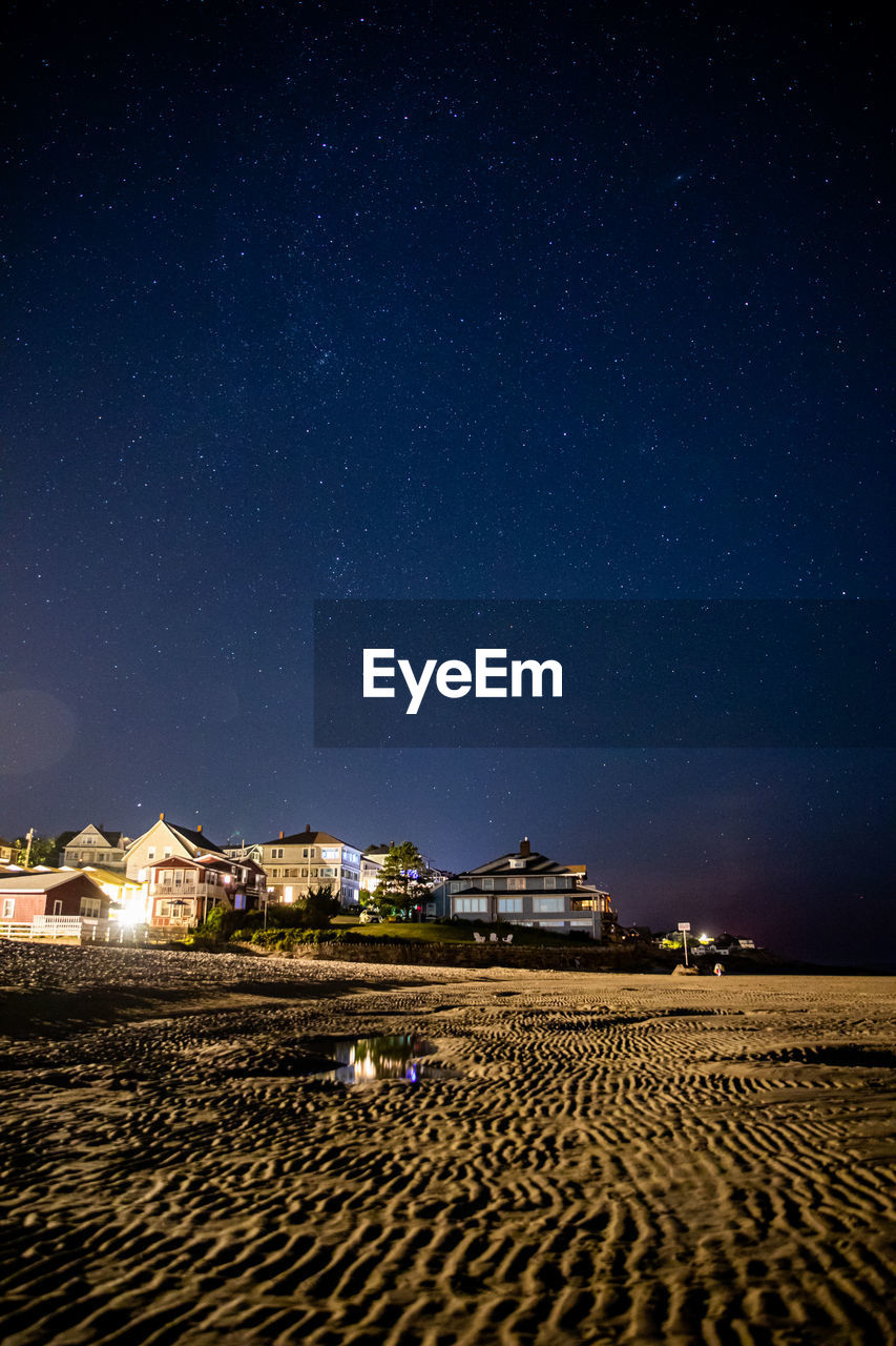 Stars in the night sky above beach houses at low tide.