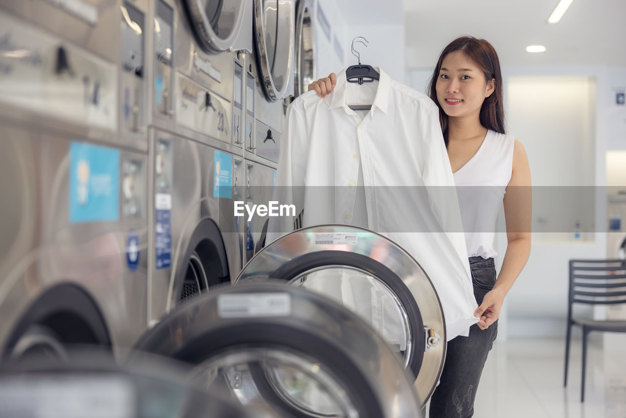 In the self-service laundry with dryer machines in the backdrop, a young woman enjoys clean ironed.