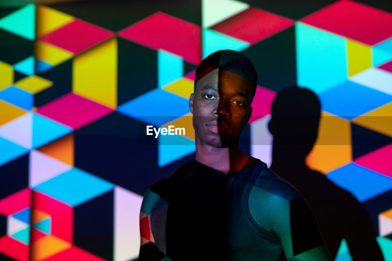 Calm black male model standing in dark studio illuminated by colorful neon light projection in shape of cubes and looking at camera