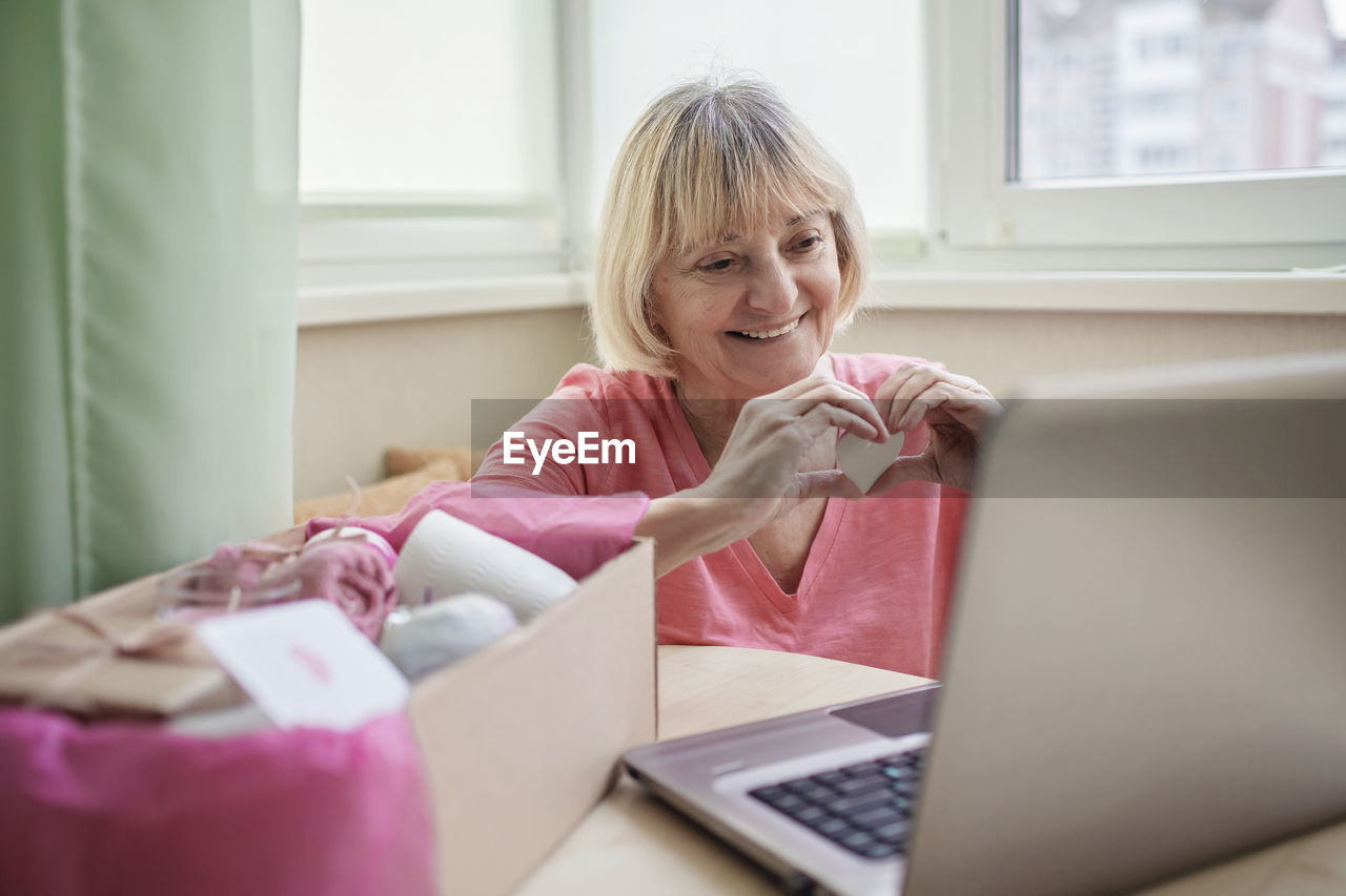 Senior woman holding heart shape using laptop at home