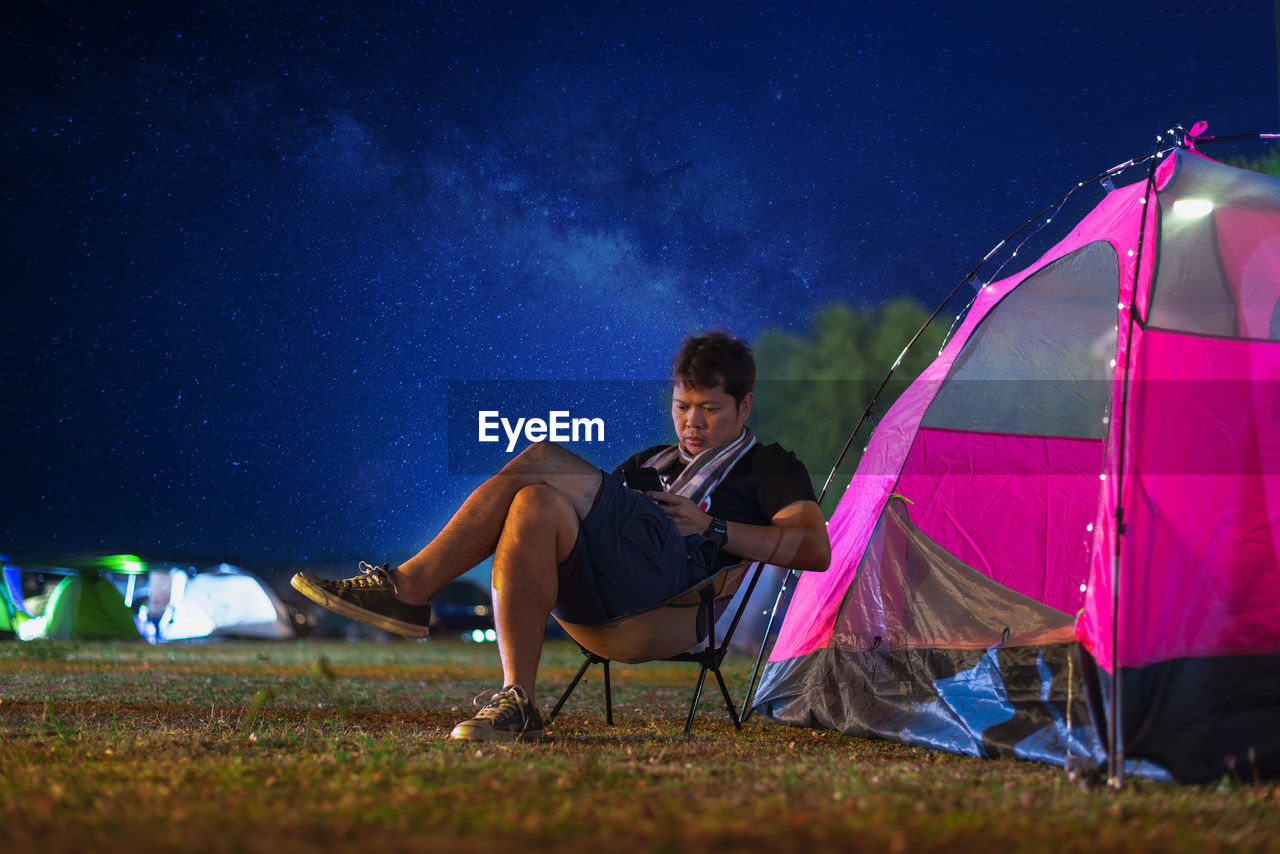 Man using phone while sitting on chair by tent on field at night