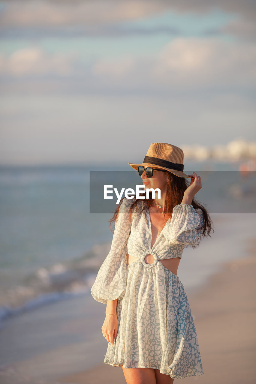 one person, clothing, adult, sea, beach, women, water, hat, land, fashion, sky, nature, young adult, standing, spring, vacation, trip, leisure activity, holiday, dress, blue, three quarter length, sun hat, casual clothing, looking, hairstyle, lifestyles, sunset, long hair, summer, focus on foreground, beauty in nature, travel, cloud, female, smiling, day, front view, person, outdoors, happiness, portrait, copy space, photo shoot, emotion, fedora, sand, relaxation, looking away, sunlight, tranquility, travel destinations, brown hair, glasses, straw hat, holding, horizon over water, tourism, carefree, enjoyment, horizon, sunglasses