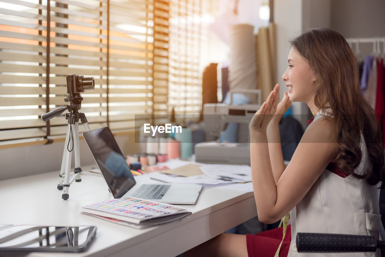 Waving her hand, a fashion designer sits at her workstation in her studio, video call.