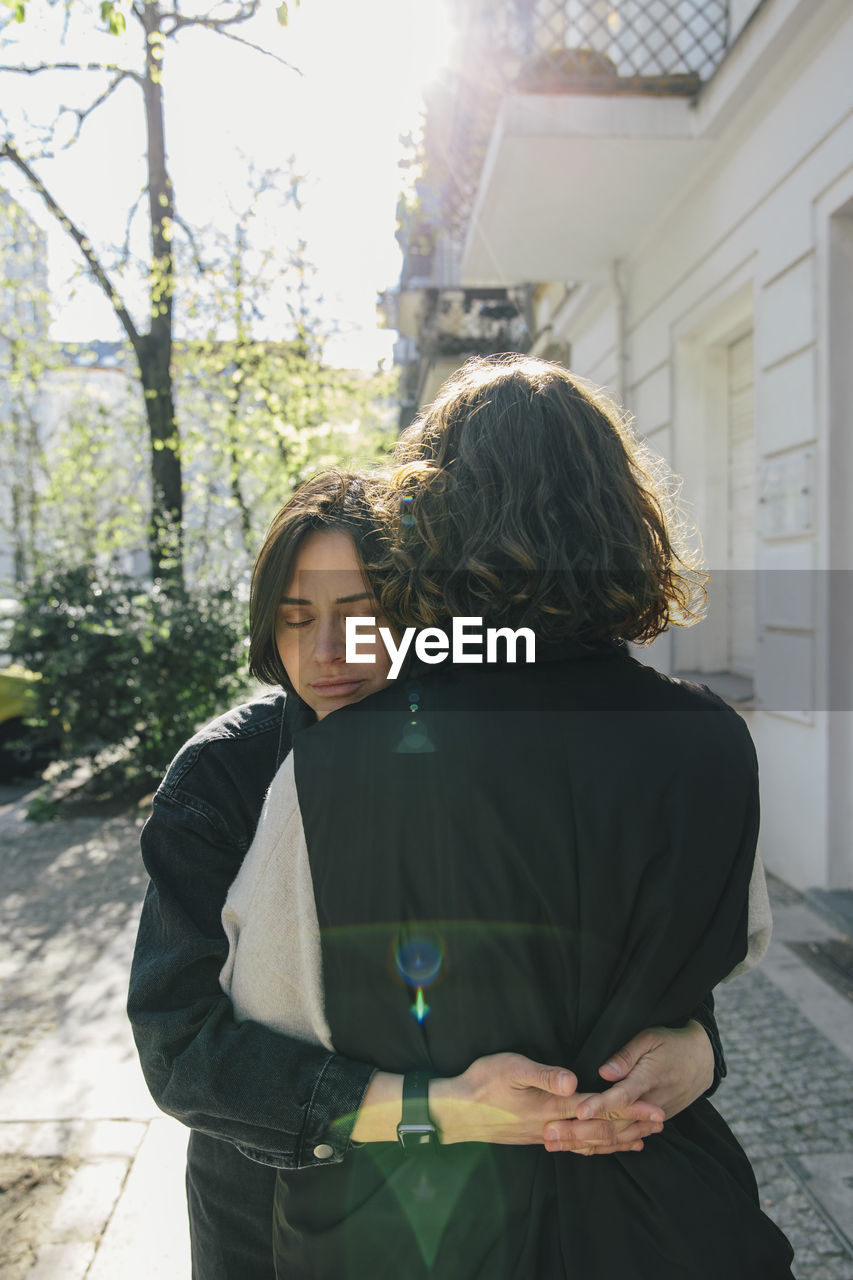 Lesbian couple embracing while standing on footpath during sunny day