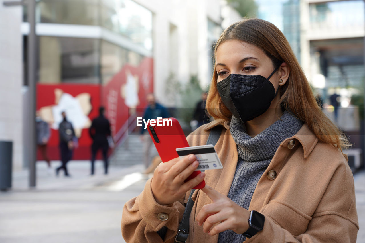 Beautiful young woman with black protective mask kn95 enters her credit card number on smart phone
