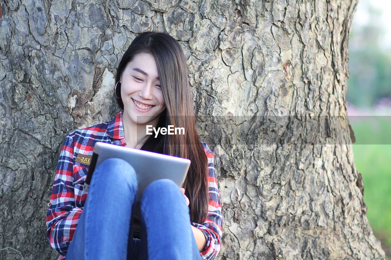 Happy beautiful woman using digital tablet sitting against tree trunk