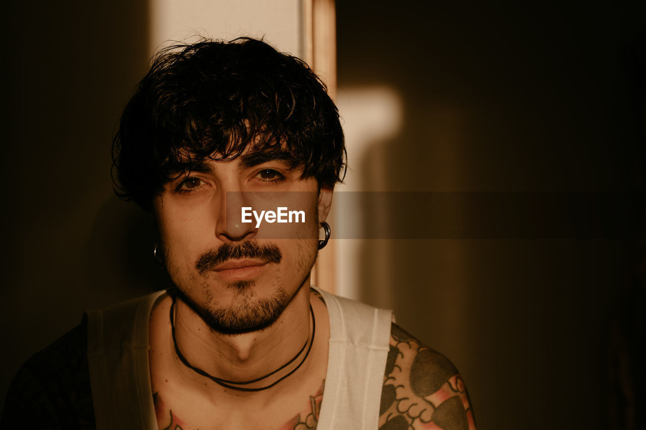 Young stylish guy with mustache and tattoos wearing white tank top and leaning against wall with back while looking at camera