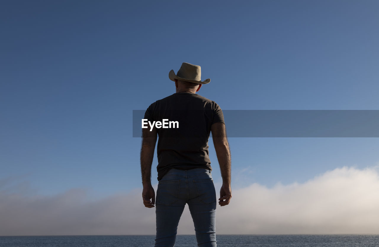 Rear view of adult man in cowboy hat and jeans against blue sky