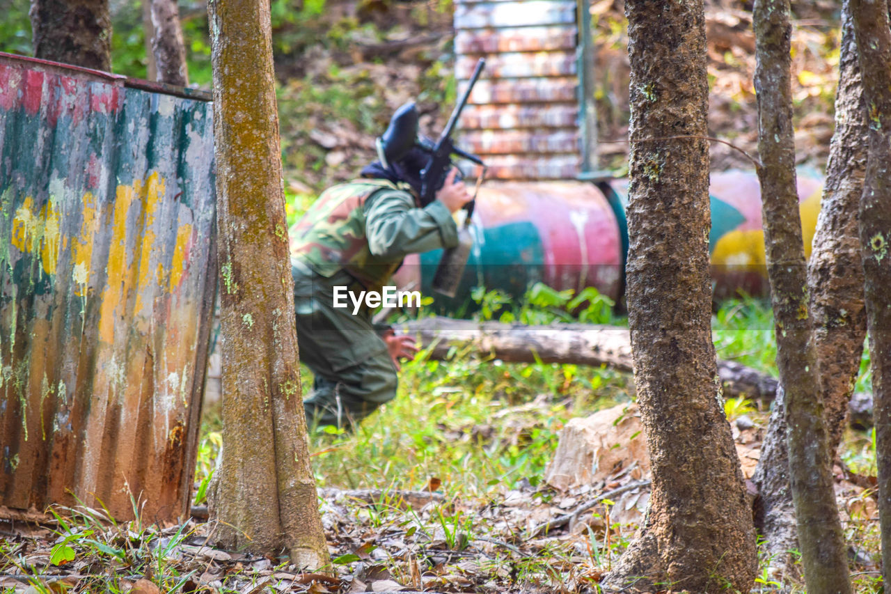paintball, shooting sport, game, sports, tree, one person, plant, recreation, men, nature, forest, day, full length, land, woodland, person, protection, leisure activity, outdoors, adult, lifestyles, tree trunk, occupation, trunk, security