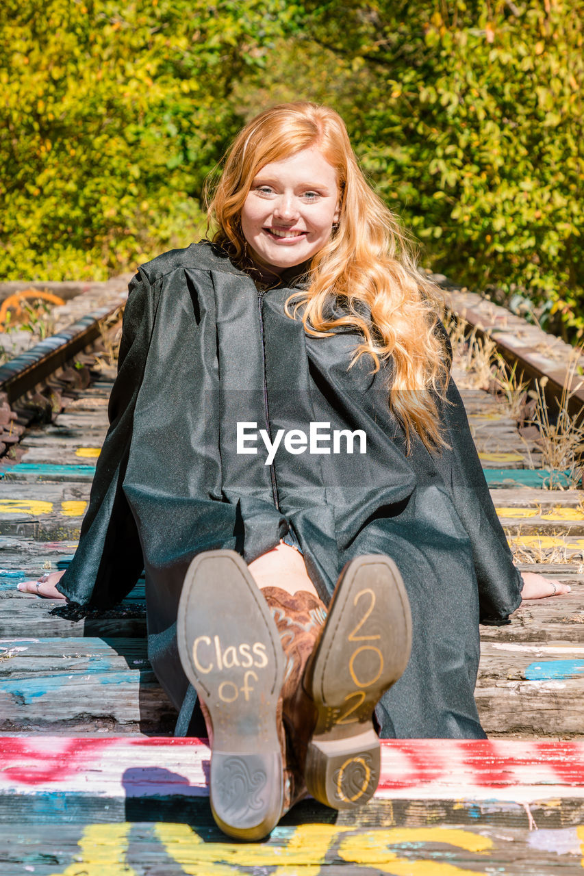 Portrait of smiling girl in graduation gown sitting on wood with text and numbers on shoes