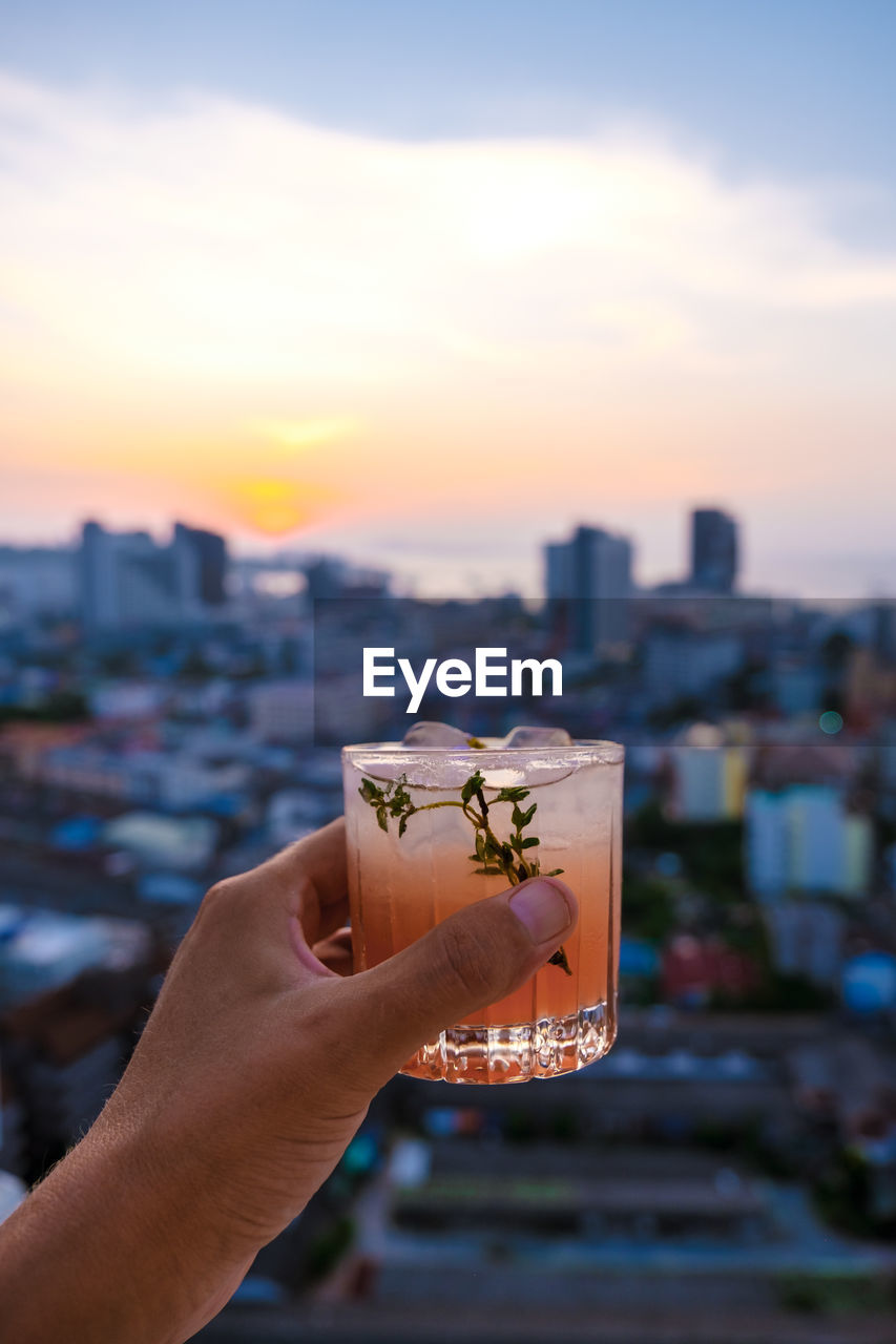 cropped hand of woman holding drink against sky during sunset