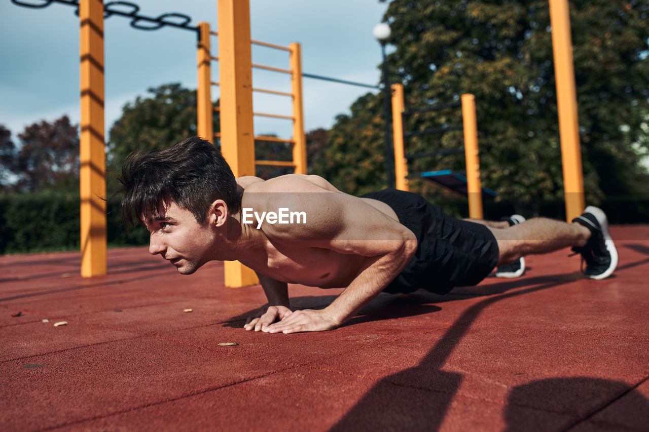 Young shirtless man bodybuilder doing push-ups on a red rubber ground during his workout