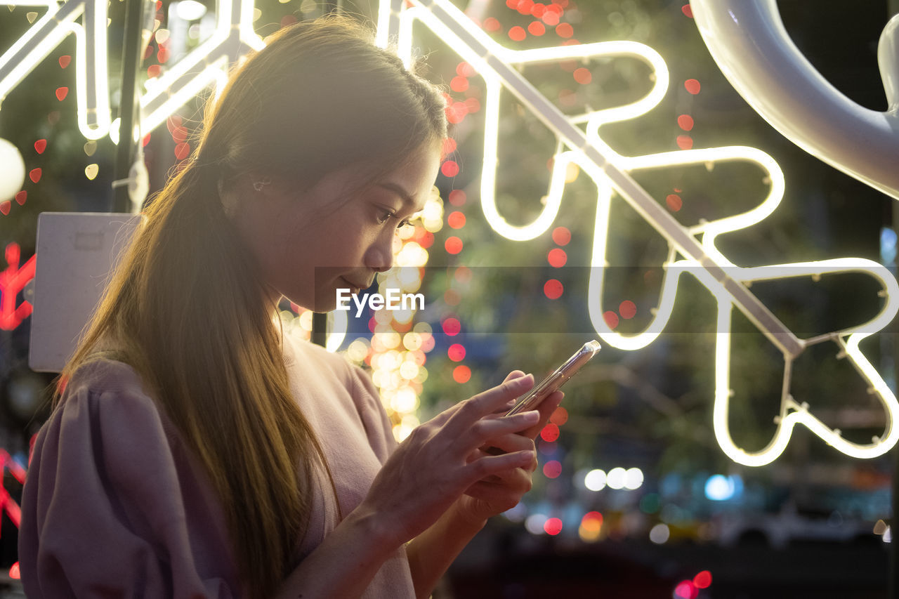 Young woman using smart phone while standing against illuminated lighting equipment