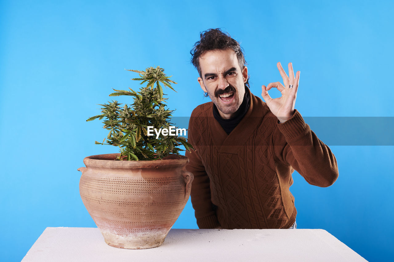 one person, flowerpot, adult, blue, men, houseplant, potted plant, plant, portrait, nature, emotion, indoors, person, smiling, growth, young adult, happiness, blue background, colored background, front view, studio shot, casual clothing, business, lifestyles, gesturing, looking at camera, standing, waist up, hand, beard, fun, facial hair