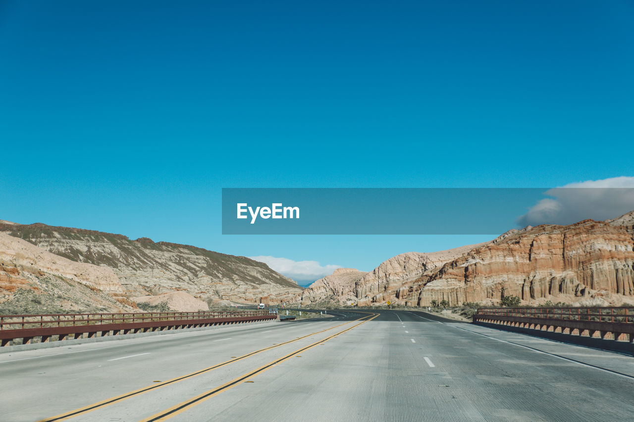 Empty road by mountains against clear blue sky