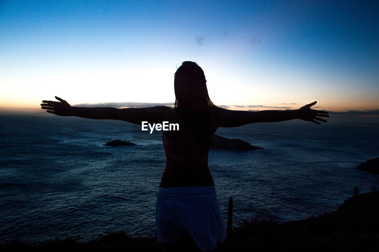 Woman with arms outstretched standing by sea against sky during sunset