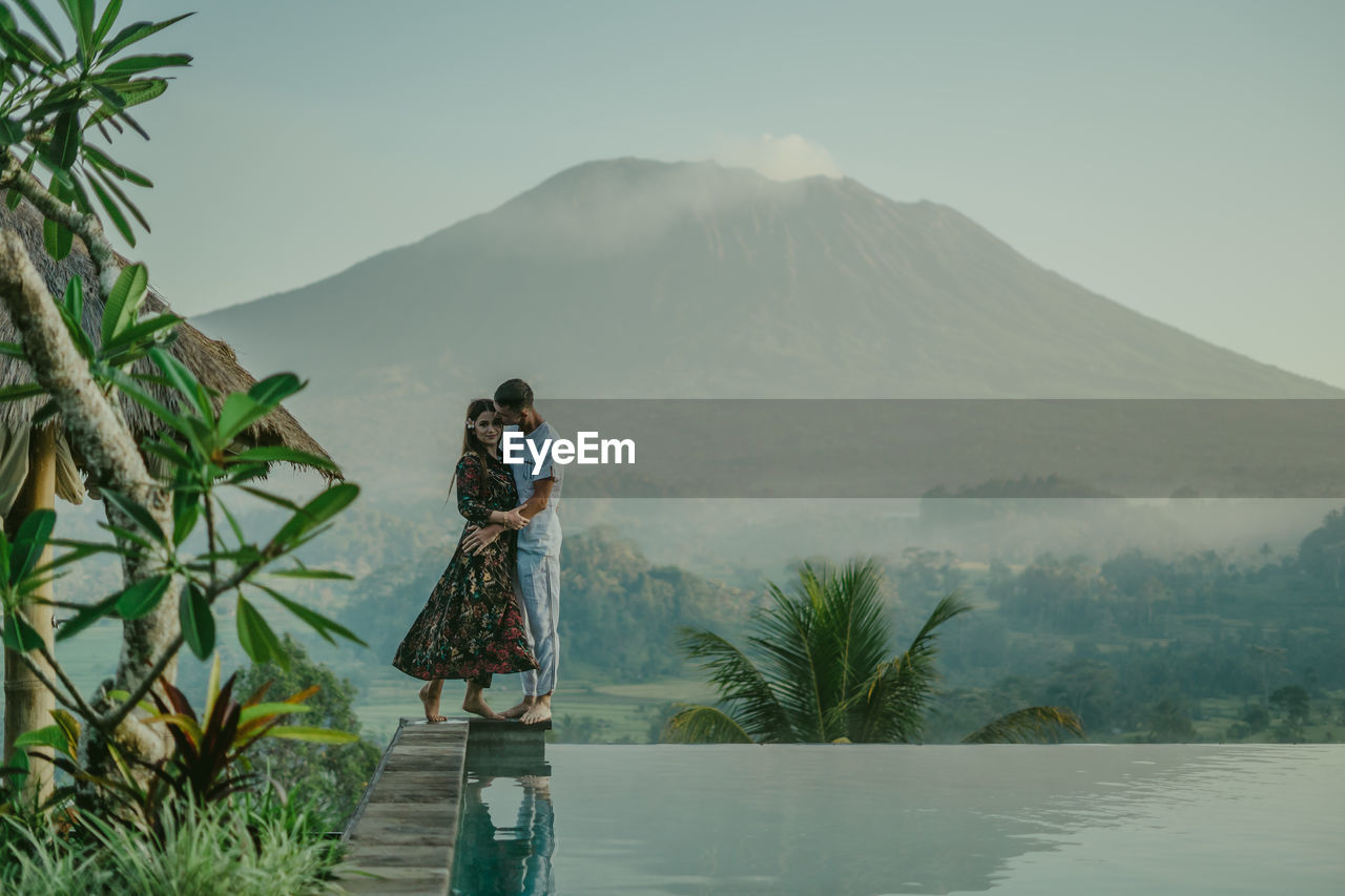 REAR VIEW OF WOMEN STANDING BY PLANTS AGAINST MOUNTAIN