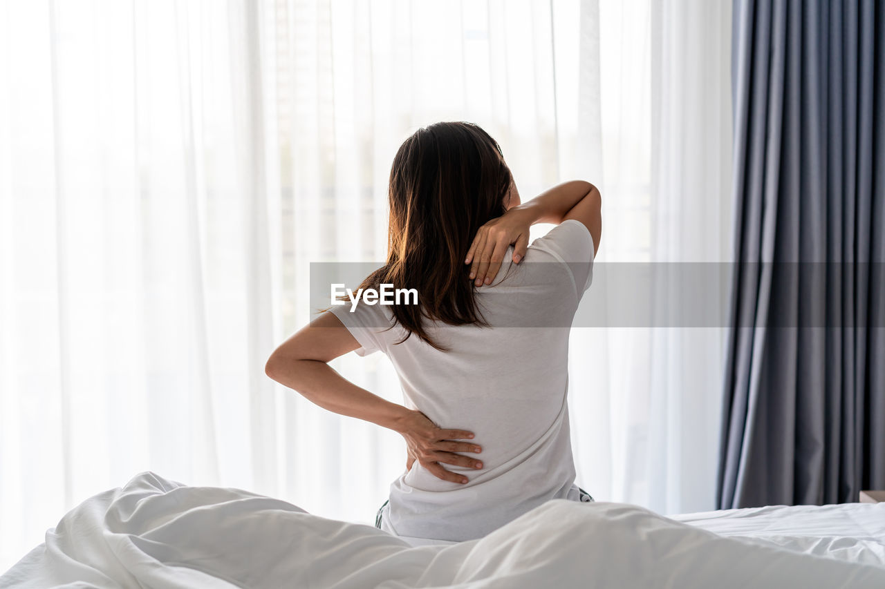 REAR VIEW OF WOMAN SITTING ON BED IN BEDROOM