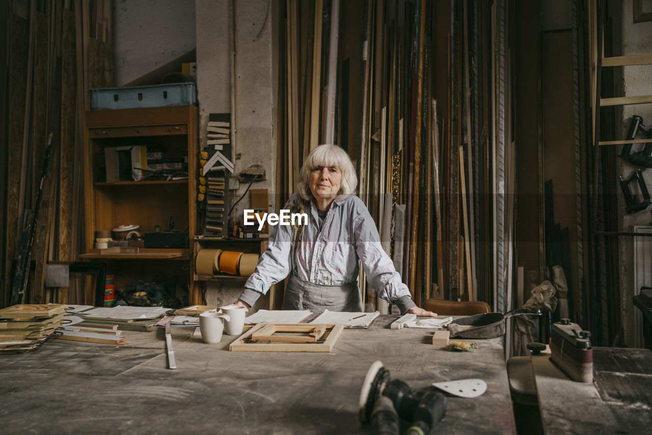 Portrait of confident senior female carpenter leaning on workbench at repair shop