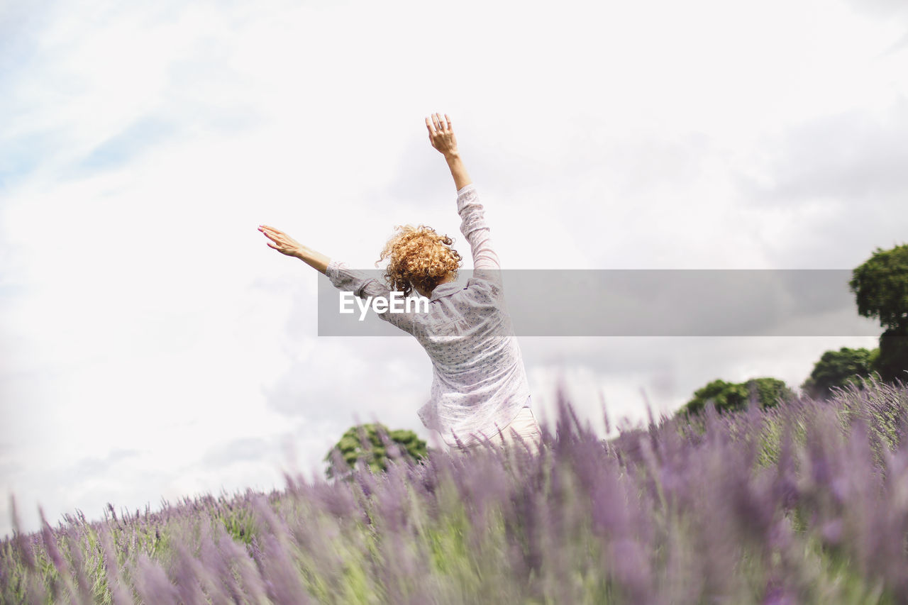 Rear view of woman with arms raised on lavender field against sky