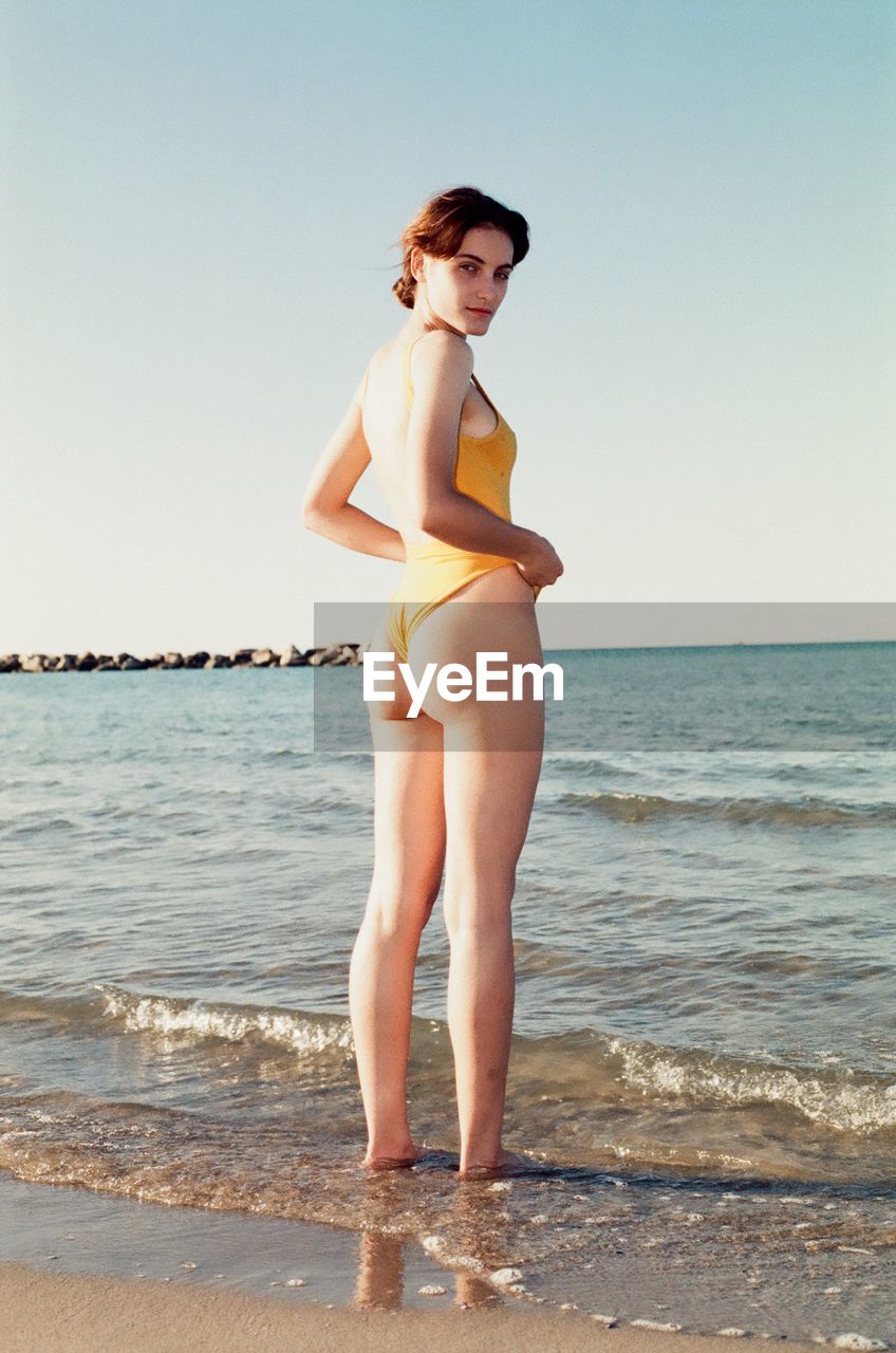 Portrait of woman in swimsuit standing on beach against clear sky