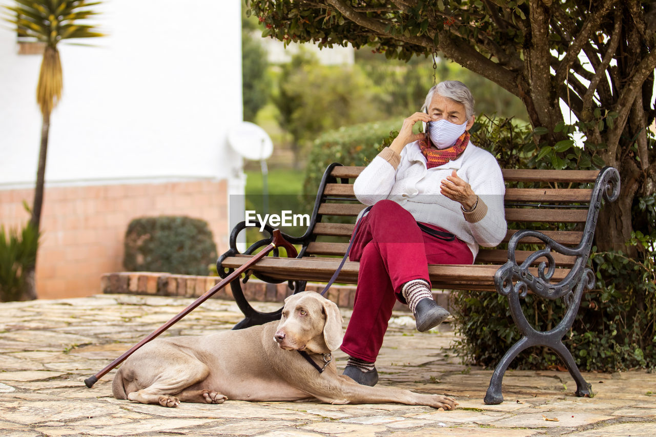 Full length of senior wearing mask with dog sitting outdoors