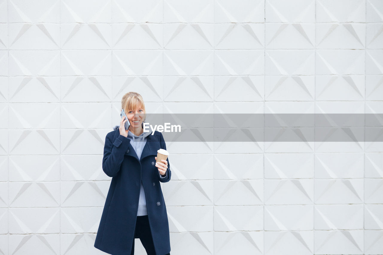 Full length of a woman using phone while standing against wall