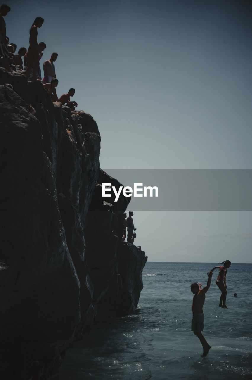 PEOPLE ON ROCKS AT BEACH AGAINST CLEAR SKY