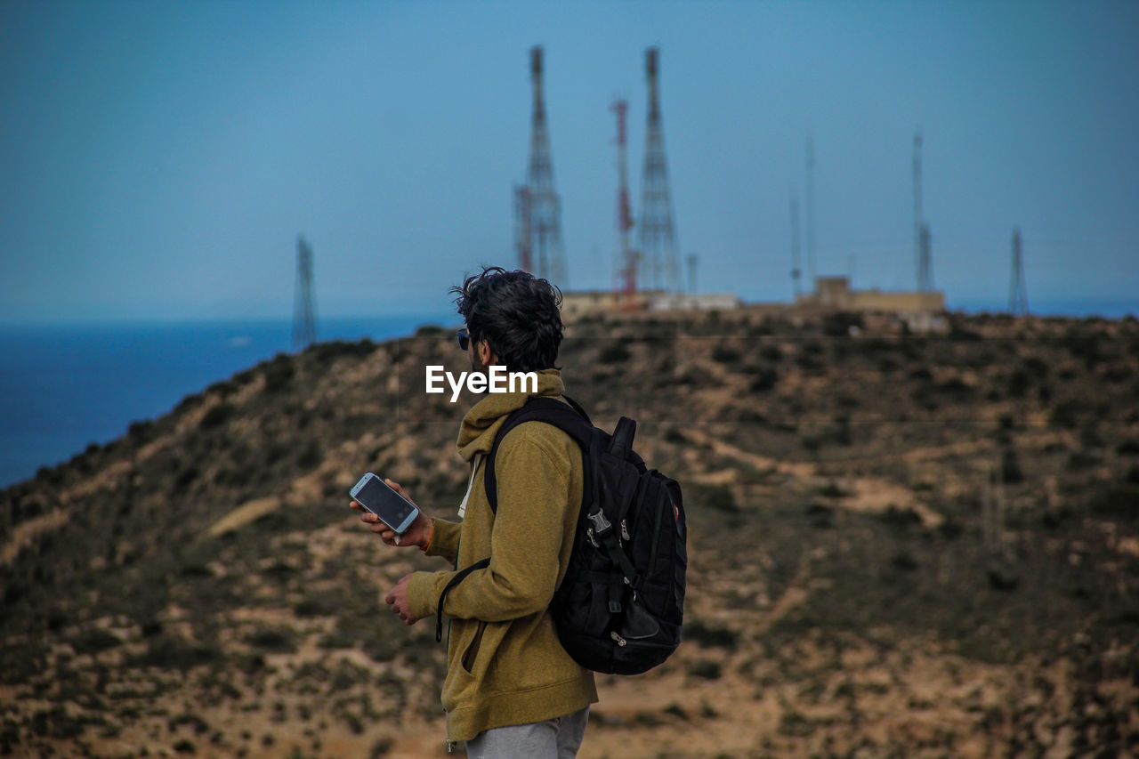 REAR VIEW OF MAN USING SMART PHONE AGAINST SKY