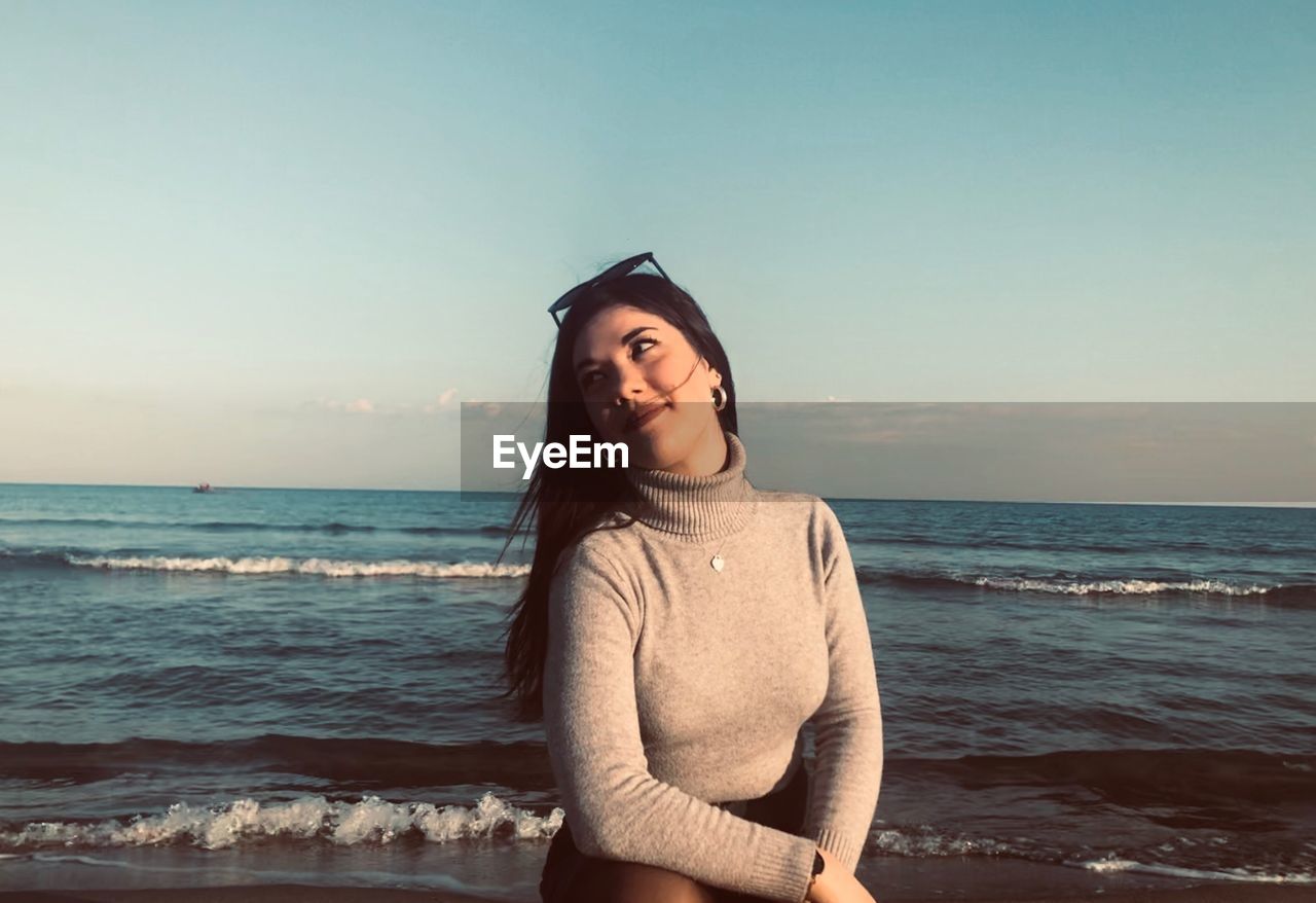 Smiling young woman sitting at beach against sky