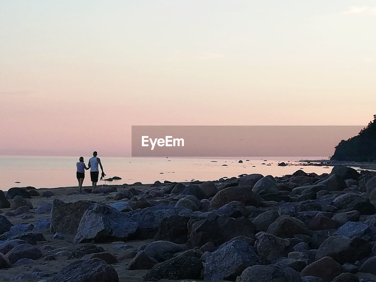 PEOPLE STANDING ON ROCK AT BEACH AGAINST SKY DURING SUNSET