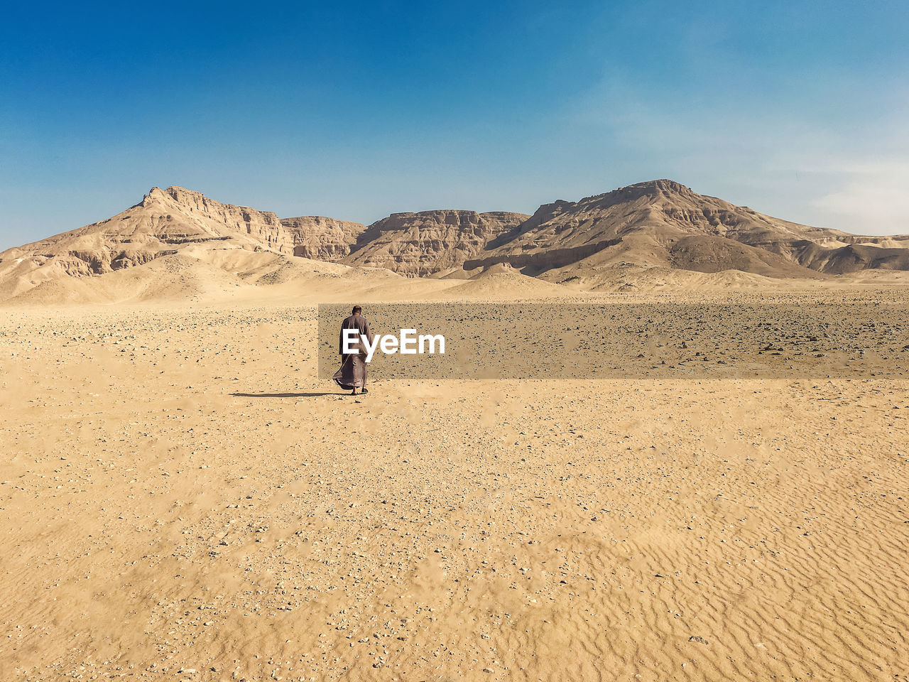 Rear view of man walking by mountains on desert against sky