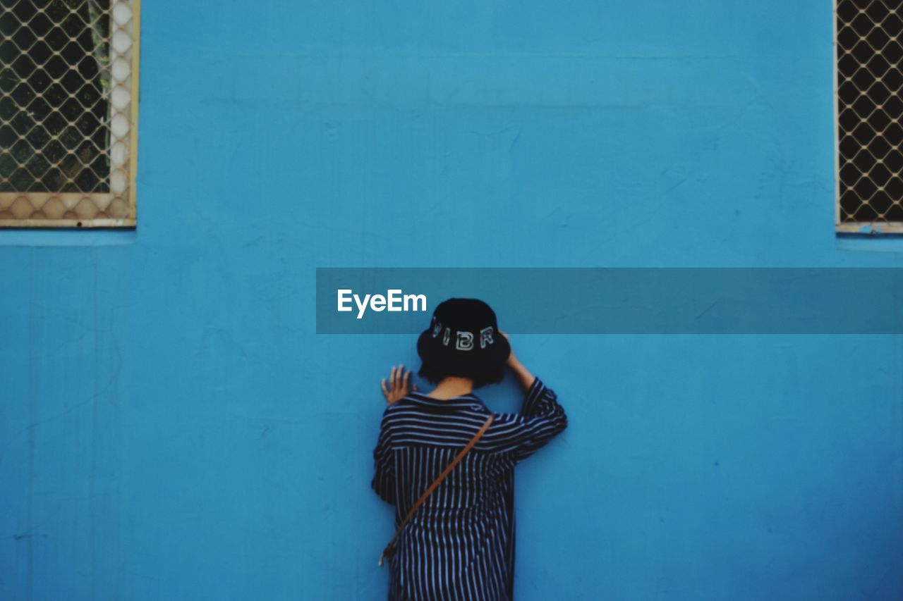 Rear view of young woman wearing hat while standing by blue wall