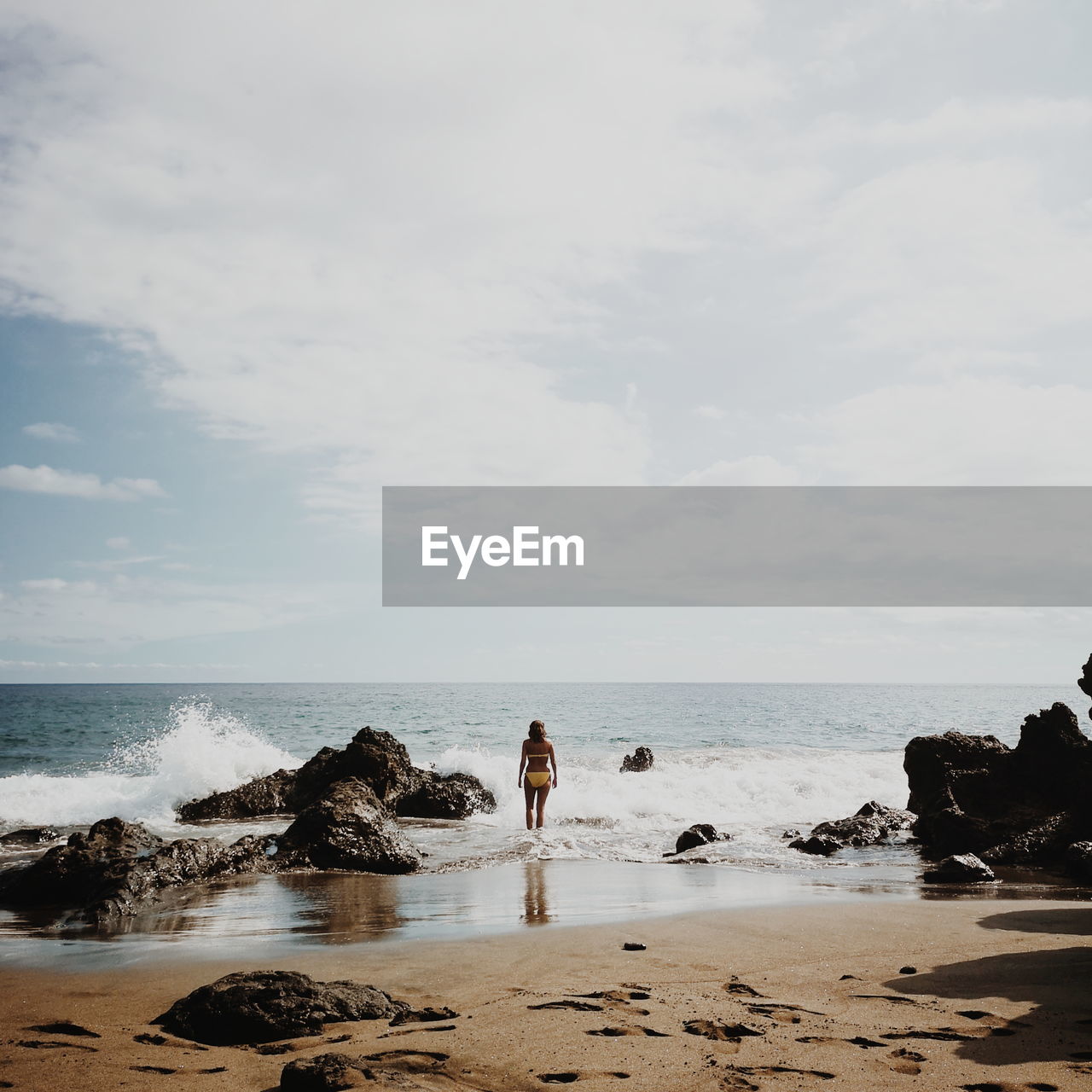 Rear view of young woman wearing bikini standing on shore at beach against sky