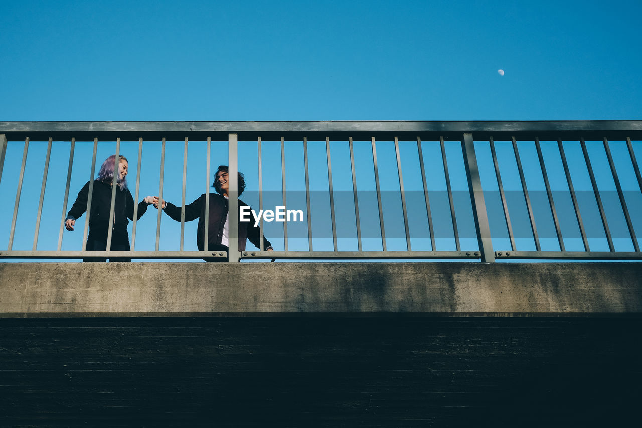 Low angle view of friends talking while standing by railing on bridge against clear sky