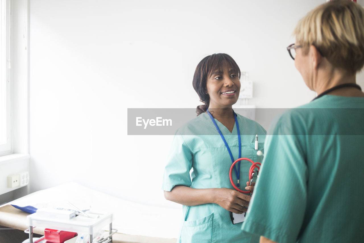 Smiling female pediatrician with bangs talking to mature coworker while standing in clinic