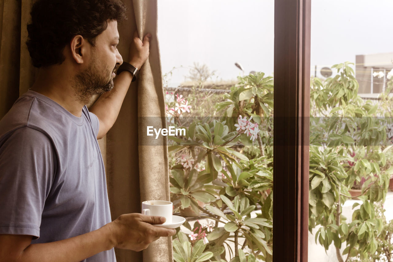 Man holding coffee cup while looking through window at home