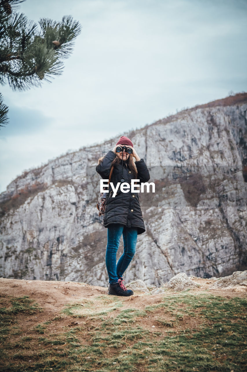 Woman looking through binoculars while standing on rock against sky