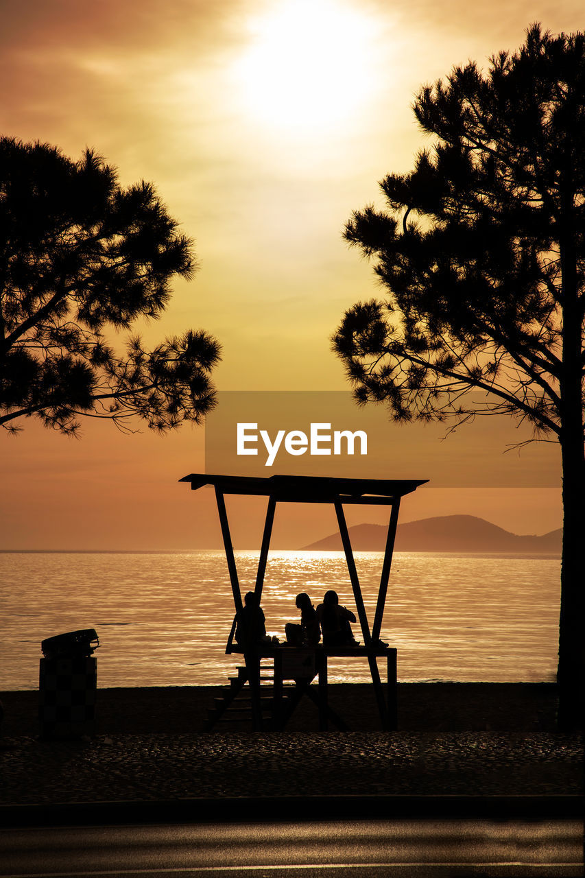 SILHOUETTE PEOPLE SITTING ON BEACH AGAINST ORANGE SKY