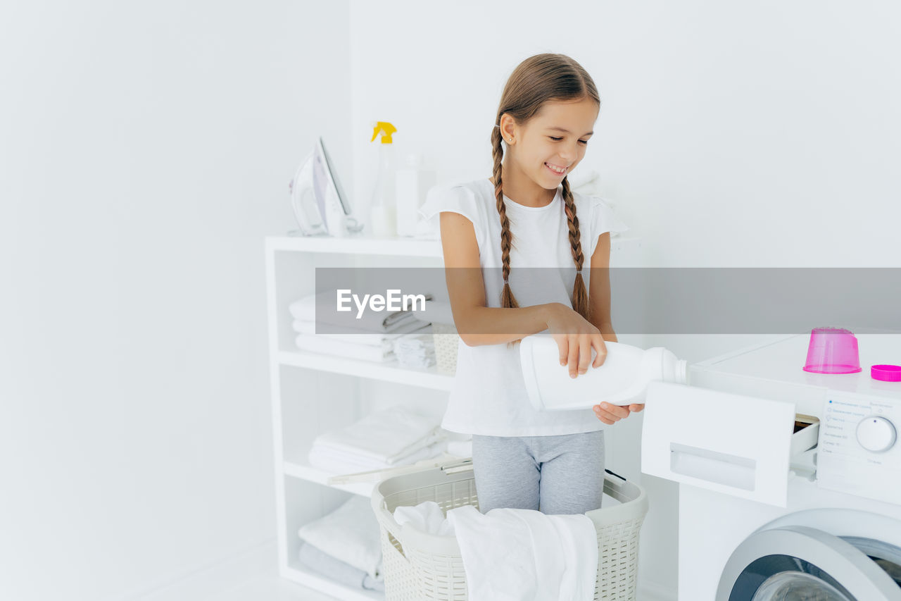 Cute girl holding detergent standing by washing machine at home