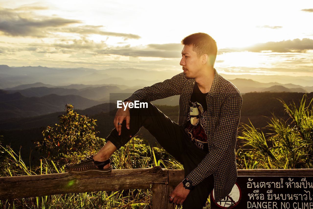 YOUNG MAN STANDING BY PLANTS AGAINST SKY