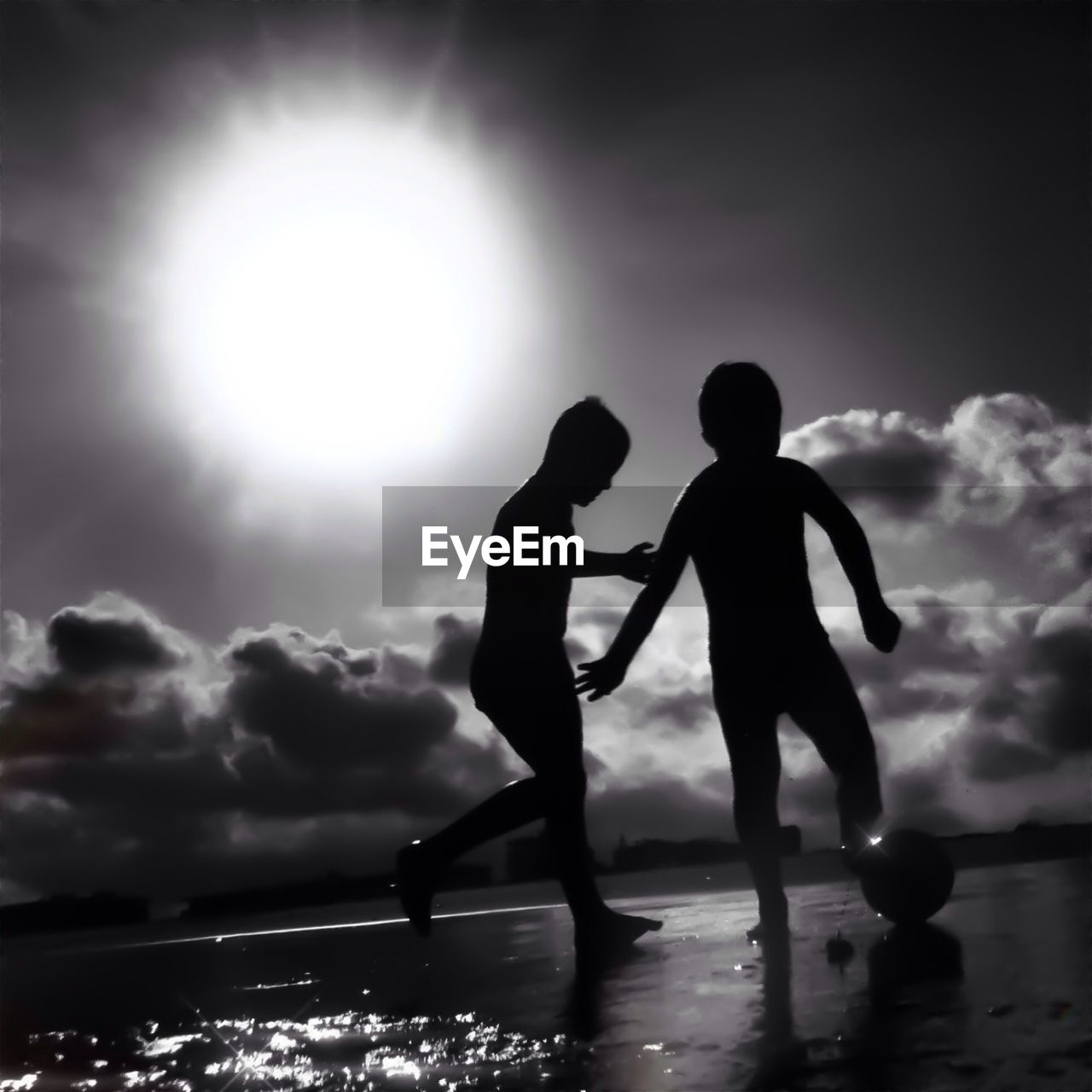 Silhouette boys playing soccer by beach against sky during sunny day